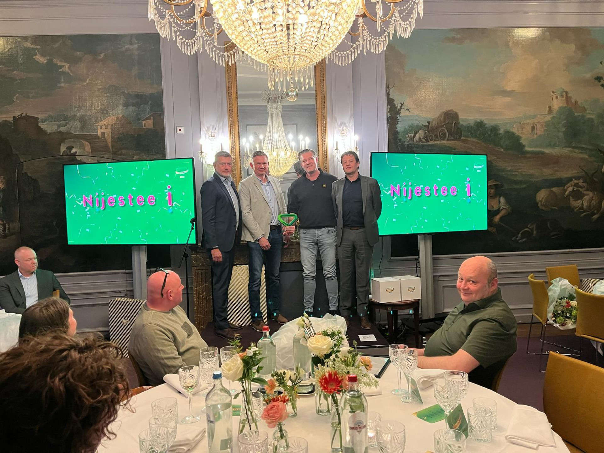 Een groep mensen poseert voor de camera tijdens een evenement met tafels en schermen op de achtergrond.